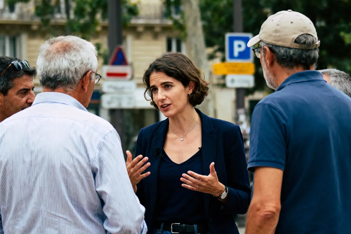 Un groupe de personnes discutant à l'extérieur, une femme au centre expliquant quelque chose, arrière-plan urbain flou.