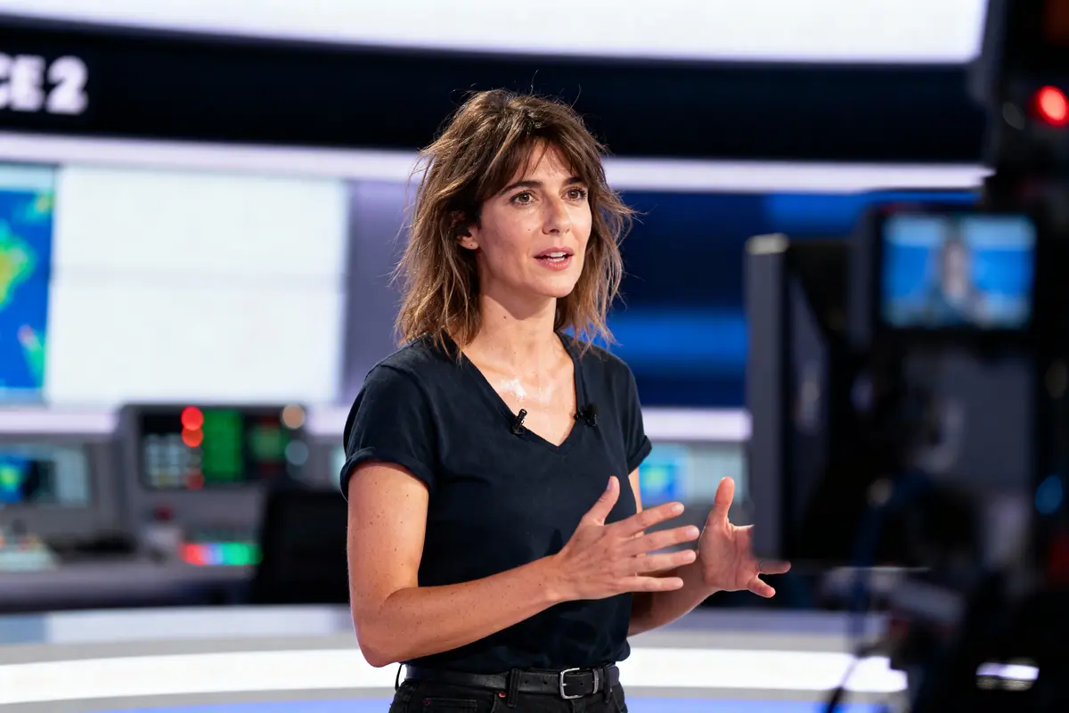 Femme sur un plateau de télévision, parlant face à la caméra, avec un fond technologique en studio.