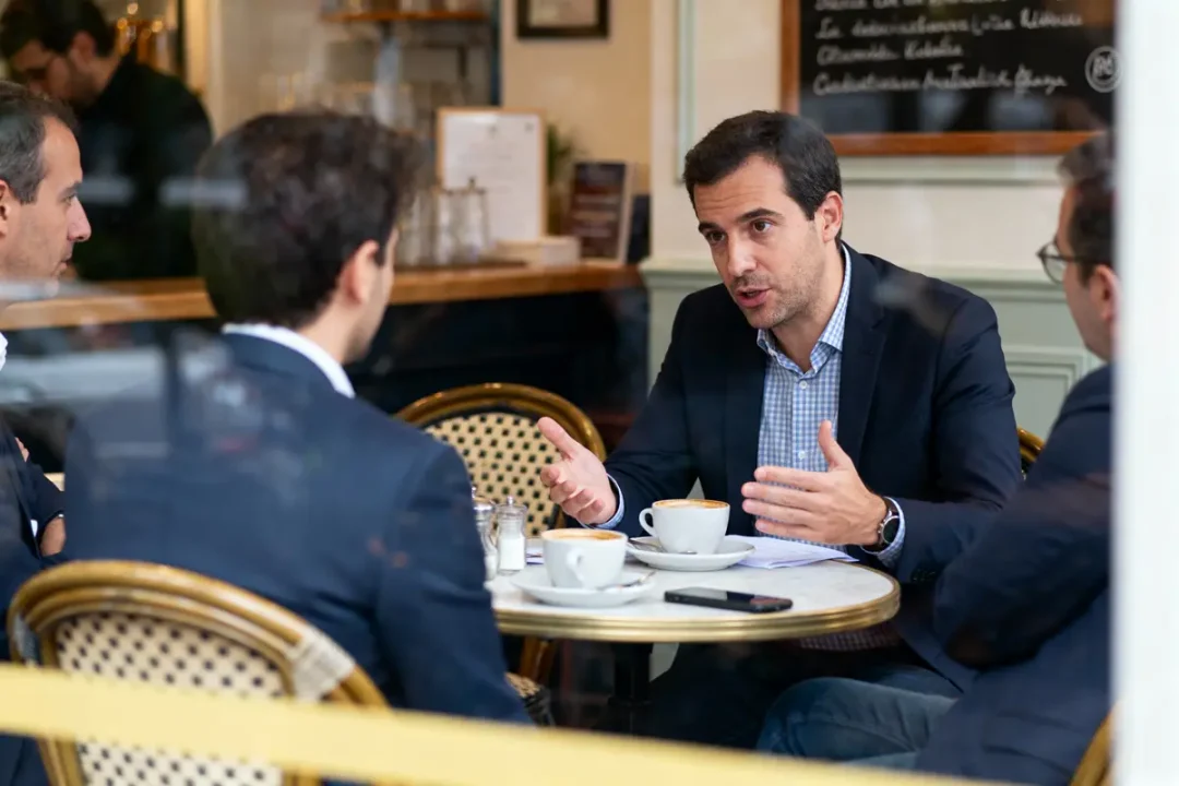 Quatre hommes discutent autour d'une table de café, avec tasses et documents, ambiance professionnelle.