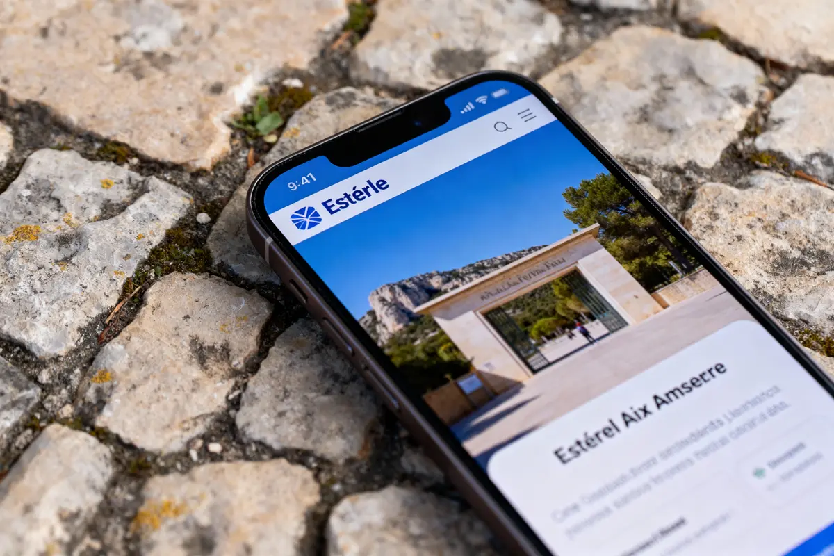 Smartphone affichant un site "Estérel" sur fond de pavés extérieurs, photo en plein air, journée ensoleillée.