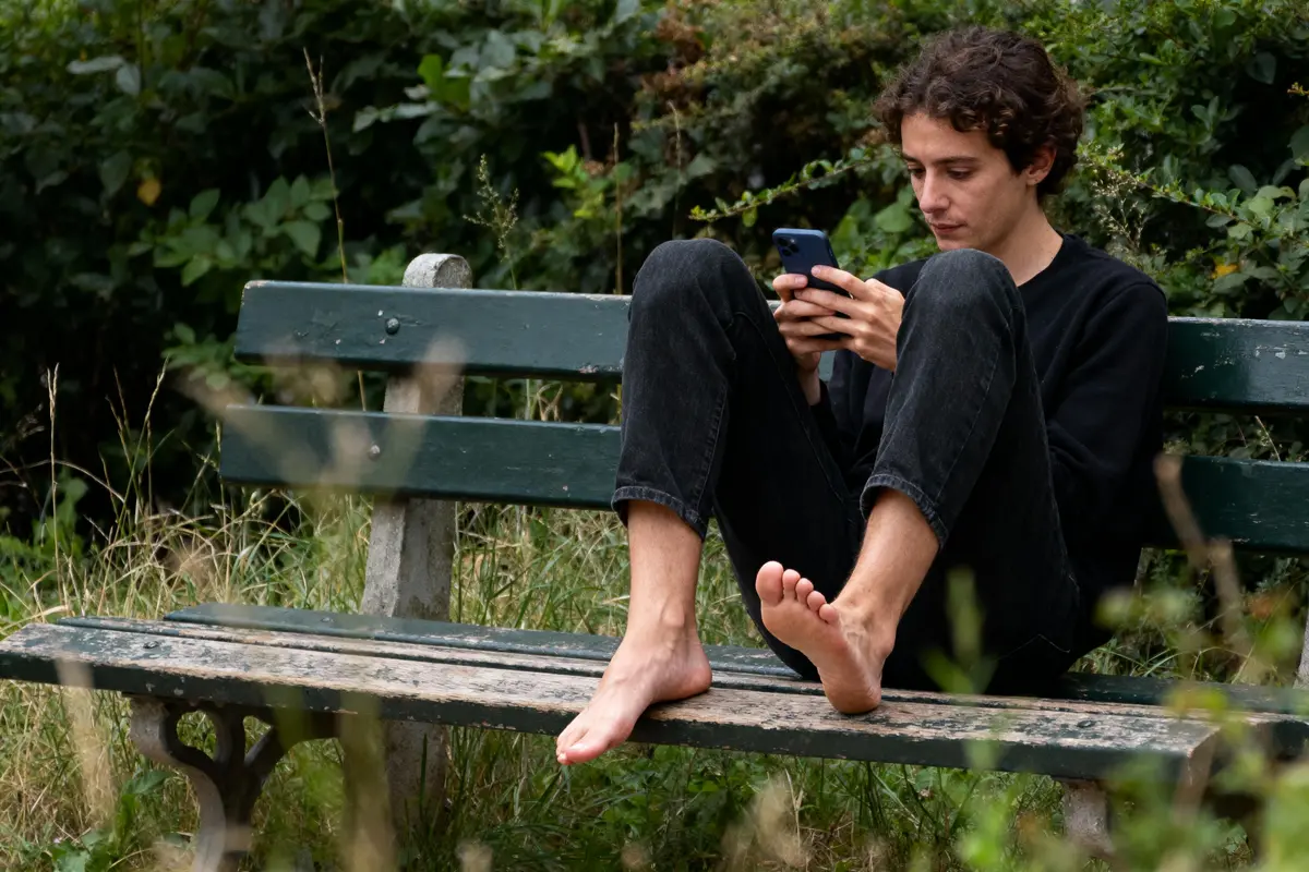 Personne assise sur un banc, pieds nus, utilisant un téléphone, entourée de verdure.