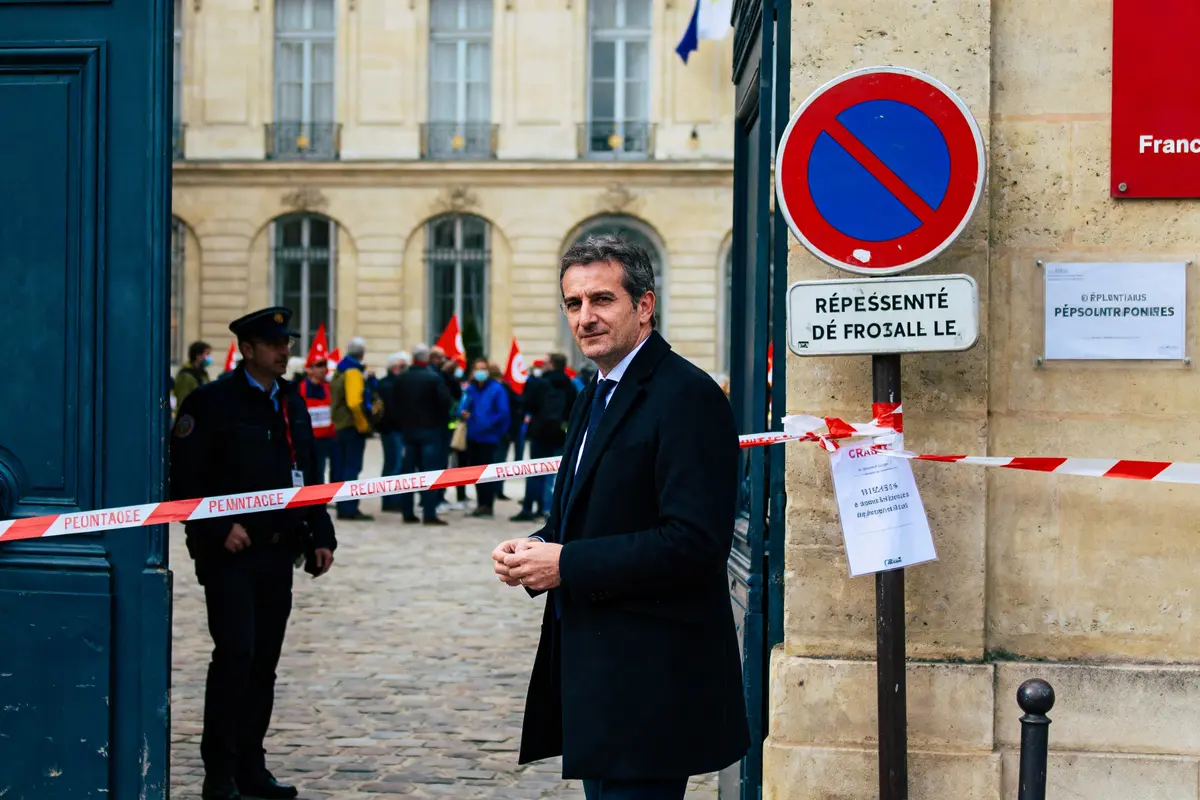 Un homme en costume debout près d'une zone sécurisée avec manifestants en arrière-plan.