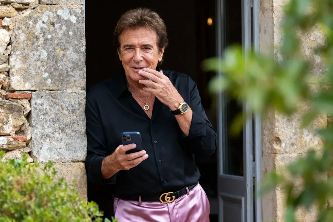 Homme souriant, en chemise noire et pantalon rose, tenant un téléphone, debout près d'une porte en pierre.