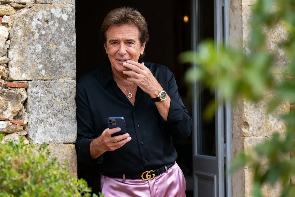 Homme souriant, en chemise noire et pantalon rose, tenant un téléphone, debout près d'une porte en pierre.