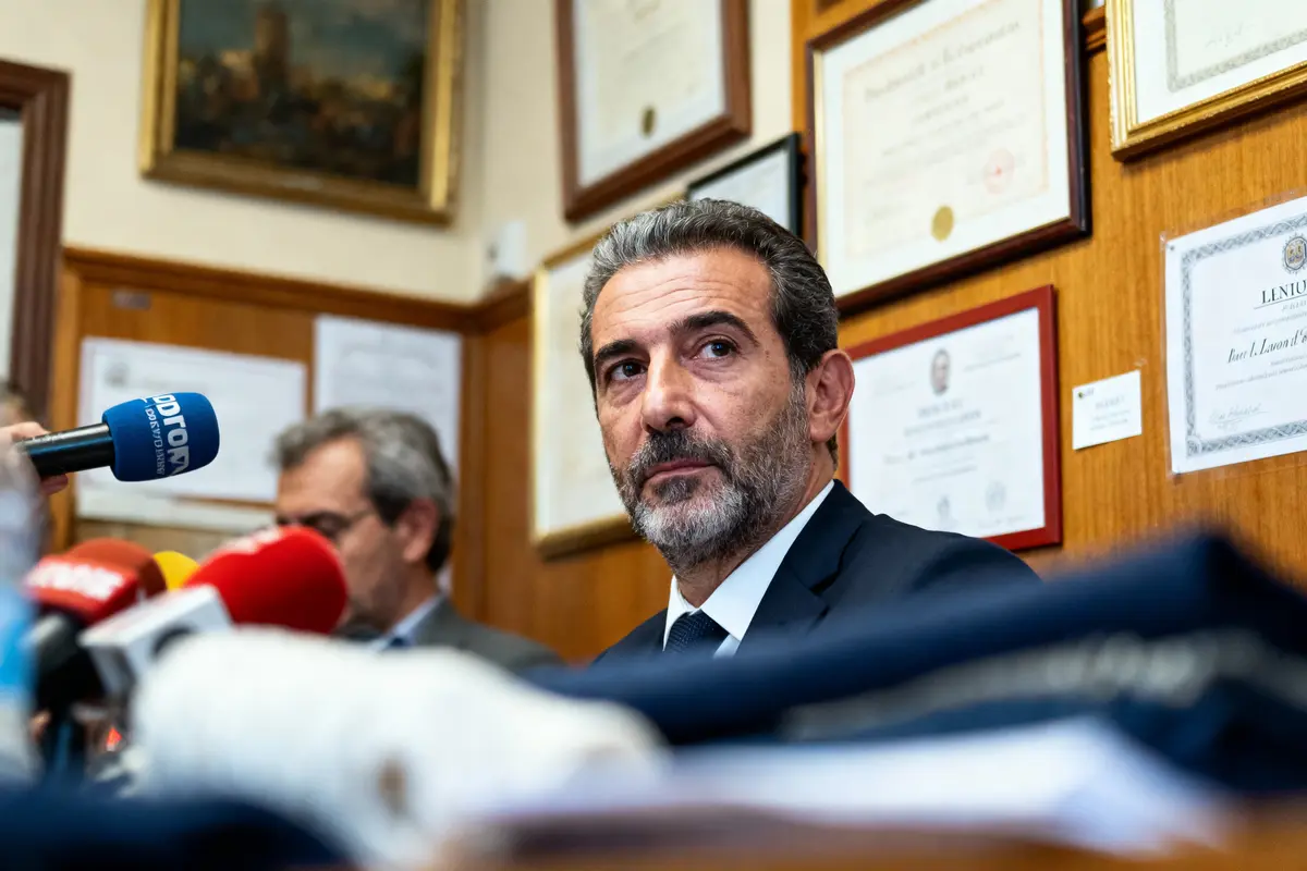 Un homme barbu en costume lors d'une conférence de presse, entouré de micros et diplômes au mur.