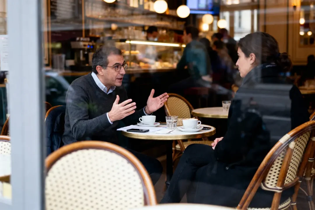 Deux personnes discutent en buvant du café à une terrasse de café, entourées d'autres clients.