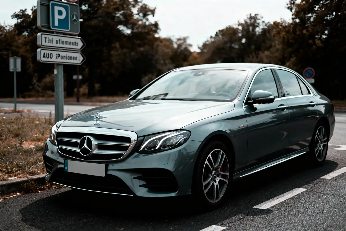 Voiture Mercedes grise garée sur la route, panneaux indicateurs en arrière-plan, arbres environnants.