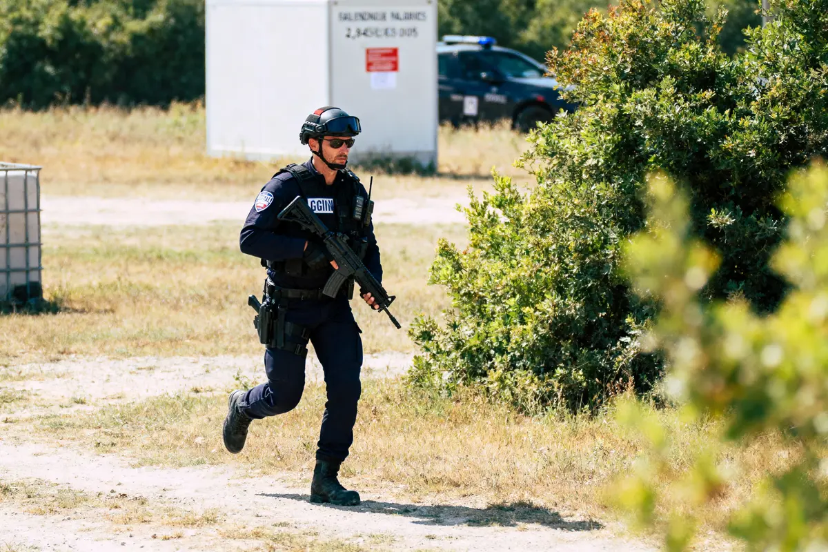 Un gendarme armé court en extérieur, portant uniforme et équipement tactique, près de buissons et véhicule de police.