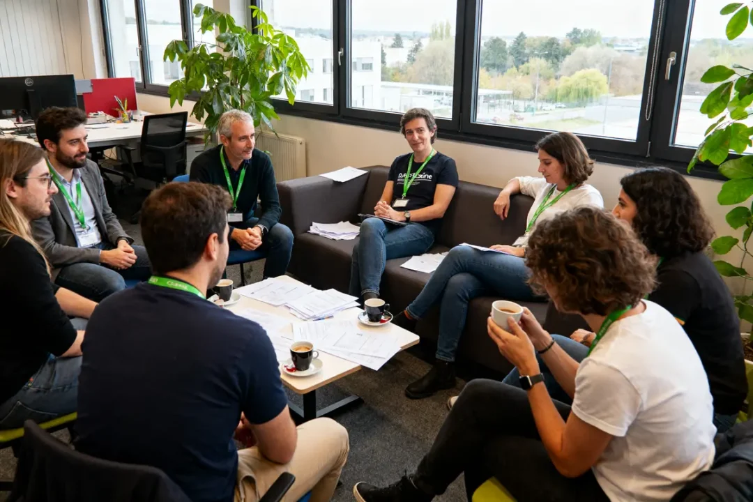 Des personnes participent à une réunion informelle dans un bureau, discutant autour de documents et de tasses de café.
