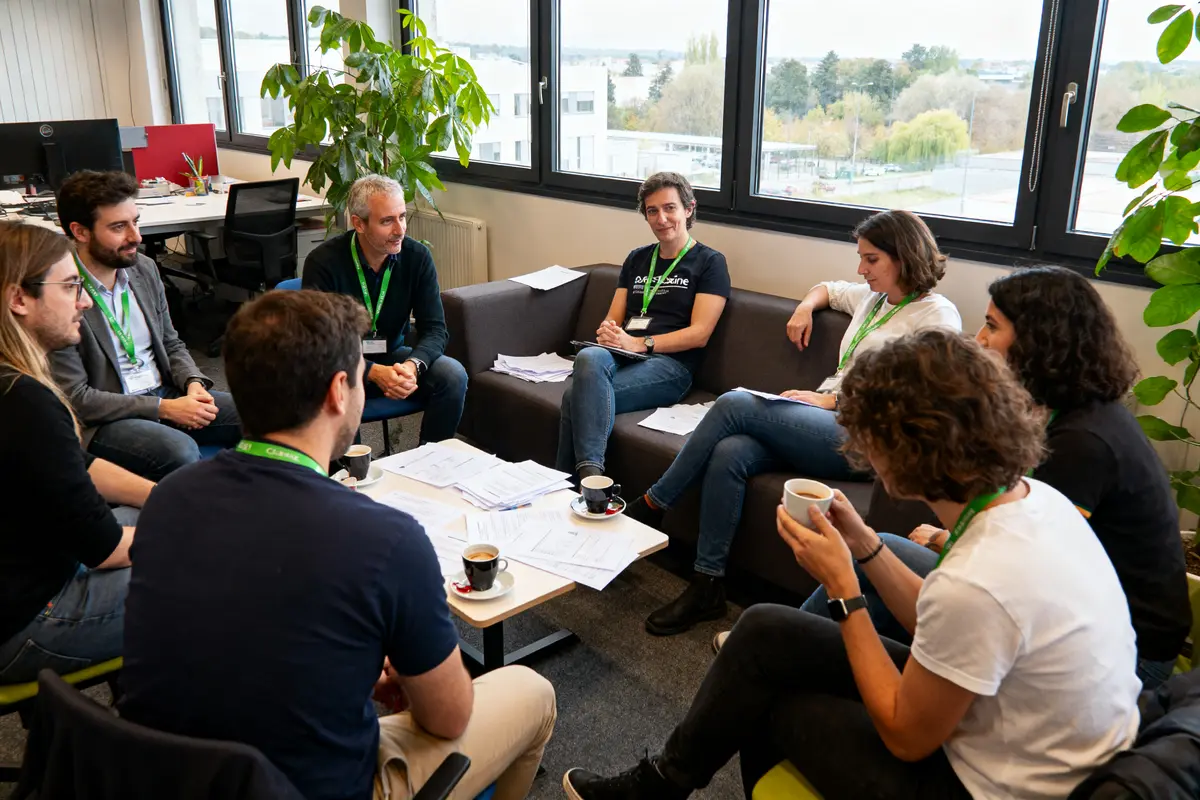 Des personnes participent à une réunion informelle dans un bureau, discutant autour de documents et de tasses de café.