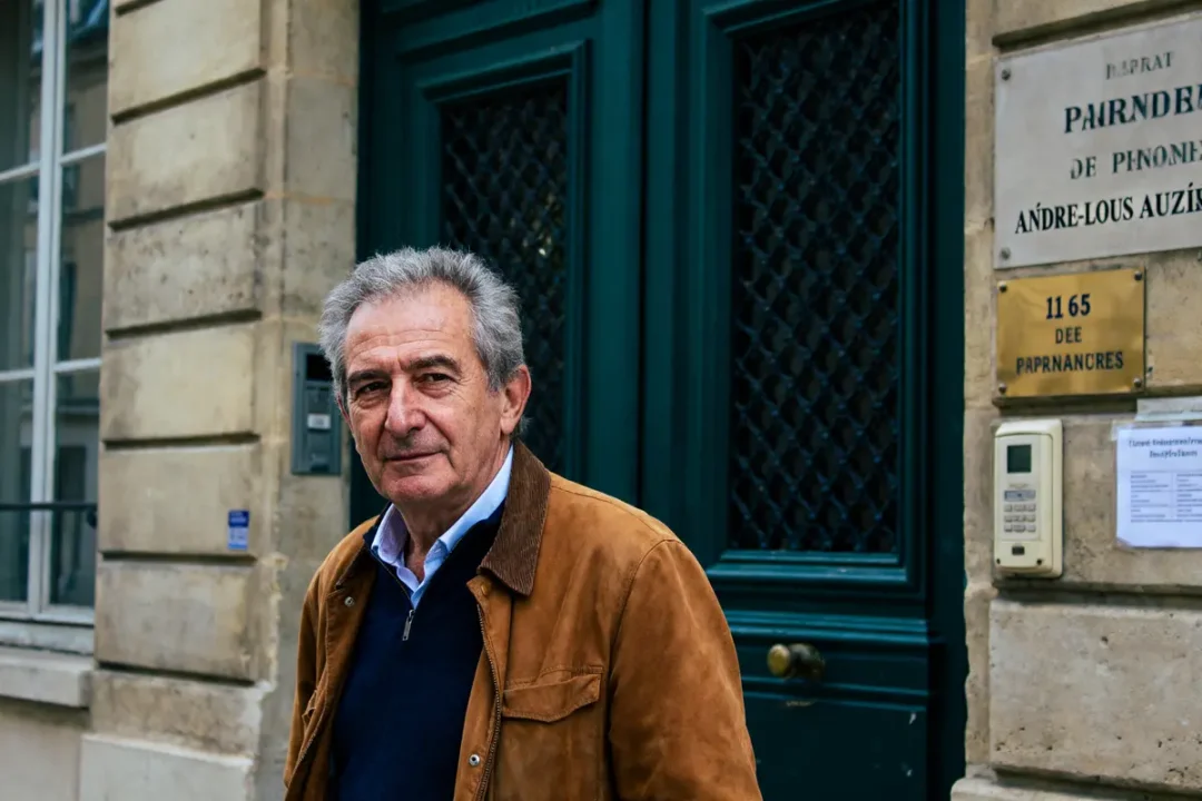 Homme en veste marron devant porte verte, mur en pierre et plaque nominative.