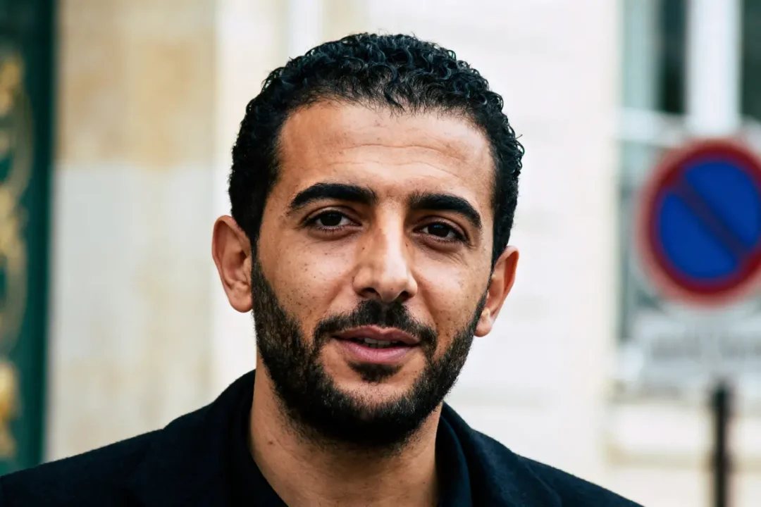 Portrait d'homme barbu avec cheveux courts, portant veste noire. Fond flou avec panneau d'interdiction visible.