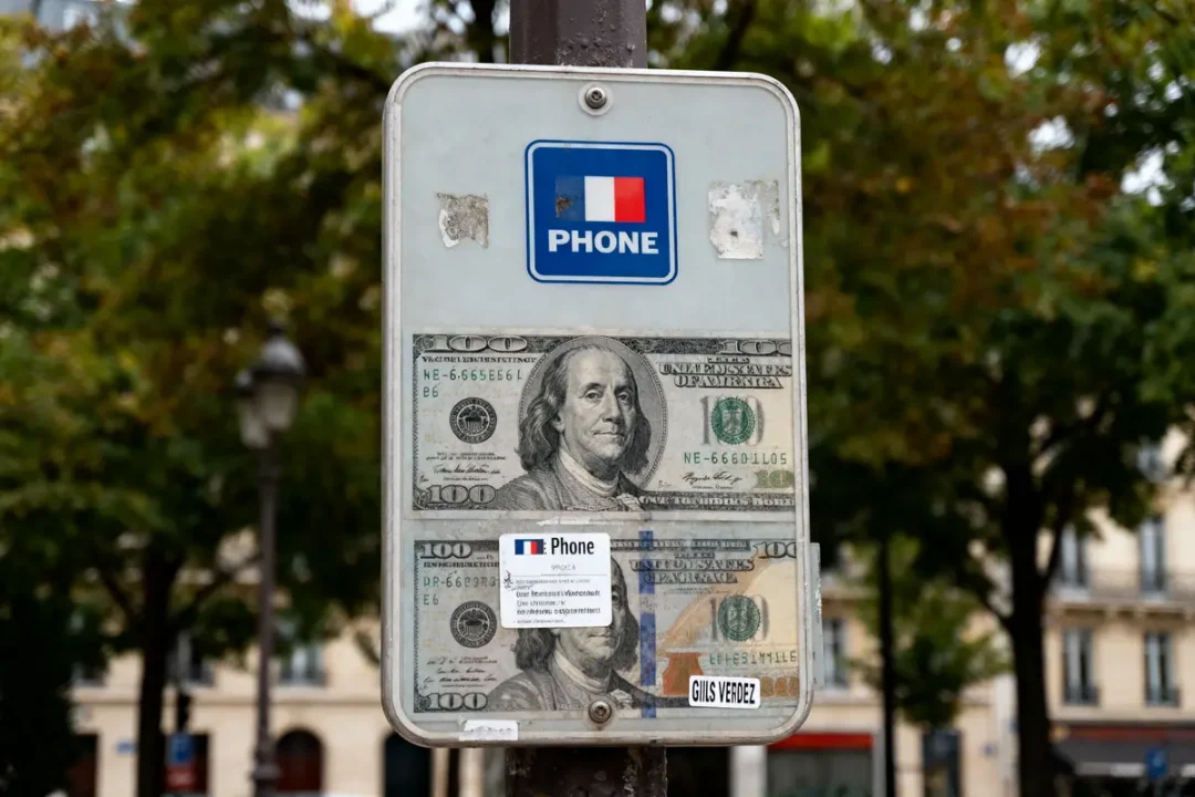 Panneau téléphonique avec collage de billets de 100 dollars, arbre et bâtiments flous en arrière-plan.