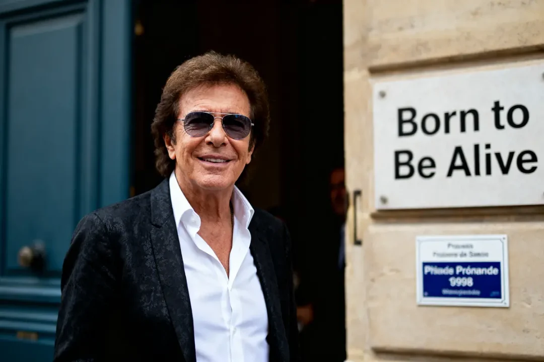 Homme en costume noir, lunettes de soleil, souriant, près d'une plaque "Born to Be Alive".