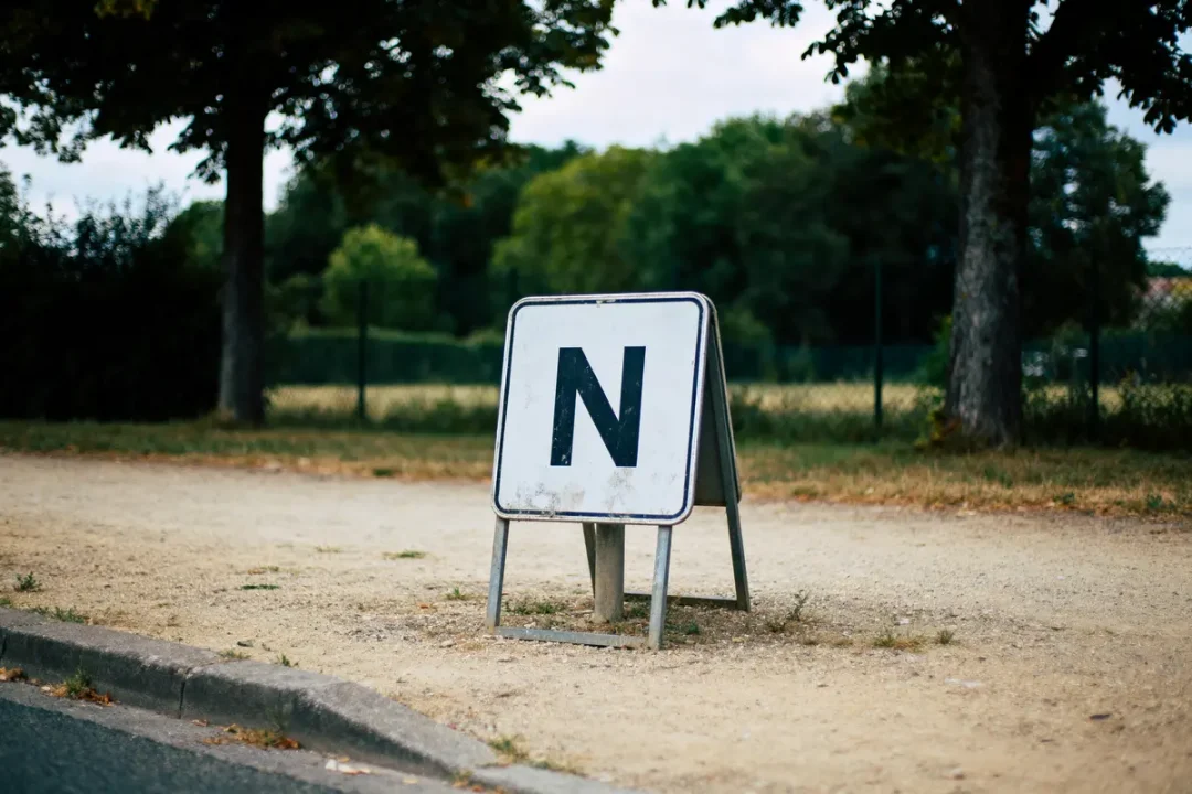 Panneau blanc avec la lettre N, sur un trottoir, arbres et herbe en arrière-plan.