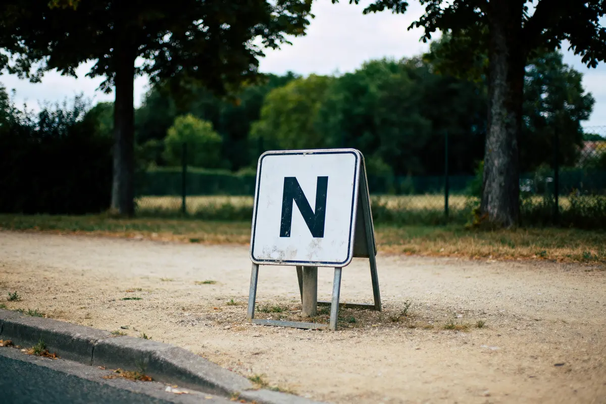Panneau blanc avec la lettre N, sur un trottoir, arbres et herbe en arrière-plan.