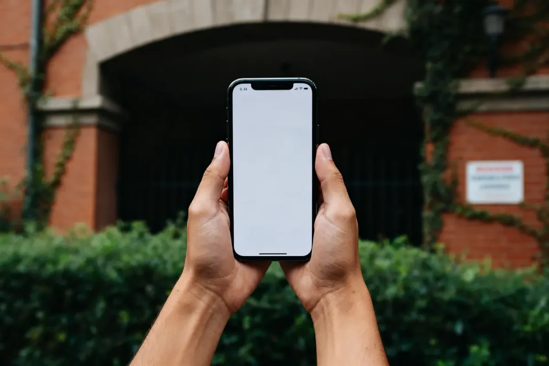 Personne tenant smartphone à écran blanc devant bâtiment en brique, bozullhuizas en arrière-plan végétal.